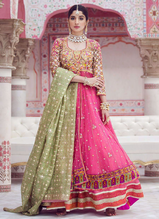 A woman models a traditional Indian anarkali gown with a pink embroidered skirt, a heavily embellished pink blouse, and a light green embroidered dupatta, posed in front of a pink architectural backdrop.