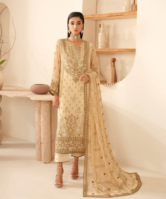 A woman models a traditional beige and gold embroidered Indian salwar kameez suit with a matching dupatta in a warm, minimalist interior setting.